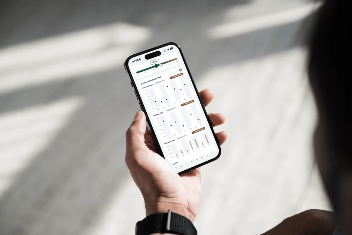Iemand houdt een telefoon vast met de gezondheidsparameters van FibriCheck open, met een hartleeftijd die 4,9 jaar jonger is en grafieken van één week hartslag in rust, ochtend-HRV en HRV-fluctuatie, telkens met het label verergering.