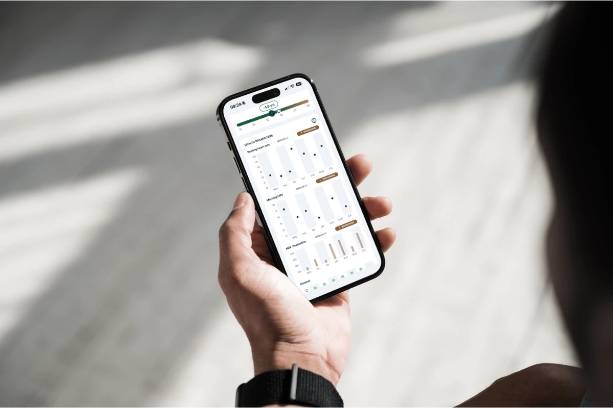 Someone holding a phone with the FibriCheck health parameters open, showing a heart age 4.9 years younger and one week of resting heart rate, morning HRV and HRV-fluctuation charts, each labeled worsening.